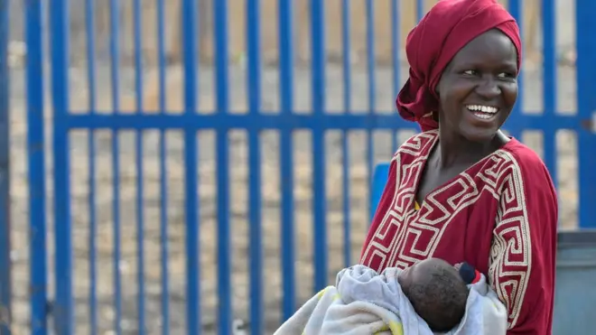 سيدة سودانية نازحة تحمل رضيعها وهي تبتسم