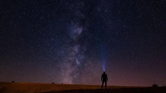 एक व्यक्ति की परछाईं, जिसने सिर पर टॉर्च पहनी है और ऊपर विशाल रात के आसमान की ओर देख रहा है, जहाँ असंख्य तारे चमक रहे हैं और बीच में आकाशगंगा (मिल्की वे) ऊपर की ओर फैली हुई है
