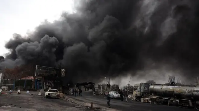 焼かれたタンクローリーなどが並ぶ奥で、巨大な黒煙が上がっている