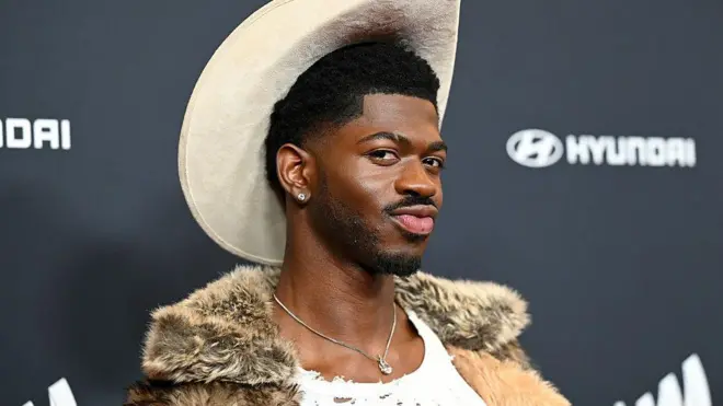 One man wit dark hair and diamond earring dey pose wit im face in front of di camera from one red carpet event. Im dey wear one beige cowboy hat, one fur coat and one silver necklace.