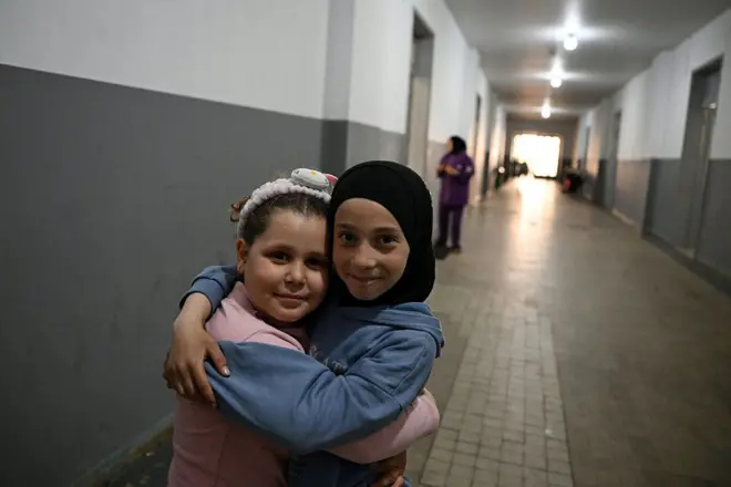 طفلتان مبتسمتان؛ إحداهما ترتدي كنزة زرقاء وحجابا أسود، والأخرى كنزة ورديّة وتضع توكة بيضاء في شعرها، وهما تتعانقان في ممرٍ طويل ممتدٍ داخل مدرسة.