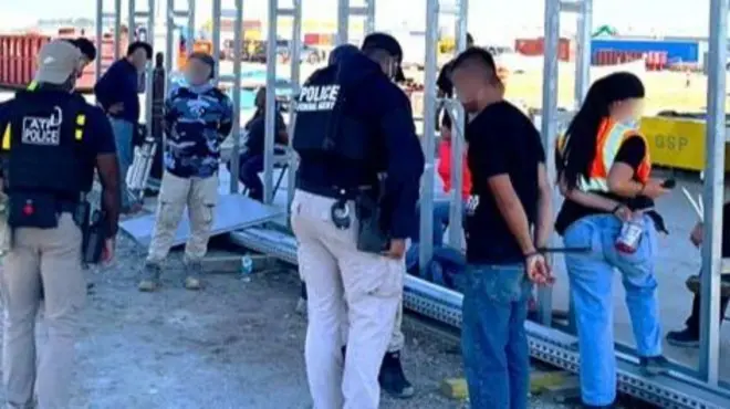 Several law enforcement agents stand near two people whose hands are bound behind their backs.