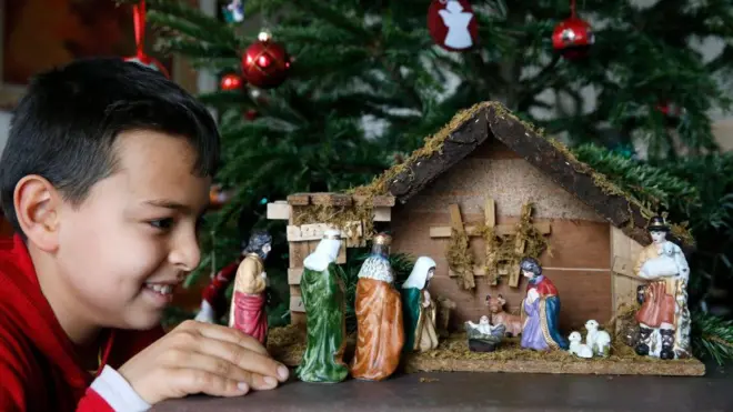 Хлопчик розглядає маленьку шопку зблизька, позаду стоїть прикрашена ялинка