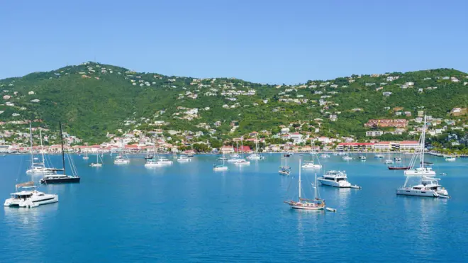 Mavi sularla çevrili bir koyda demirlemiş beyaz tekneler, önünde beyaz binalarla bezeli yeşil, ağaçlarla kaplı tepeler yer alıyor.