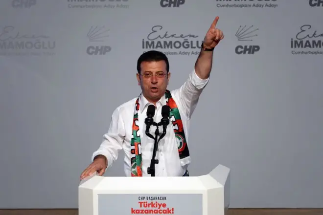Ekrem İmamoğlu bir kürsüden konuşuyor. Bir elini havaya kaldırmış. Beyaz bir gömleği var. 