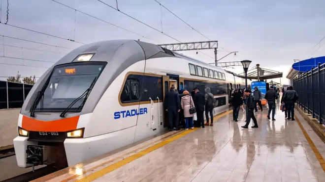 Azərbaycanın Almaniyadan aldığı Stadler markalı yeni elektrik qatarı stansiyada dayanıb və sərnişinlər yağışlı havada qatara minməyə çalışırlar.