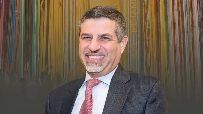 استیون فاگین با کت خاکستری و کراوات صورتی جلوی دیواری نقاشی شده با نوارهای زرد