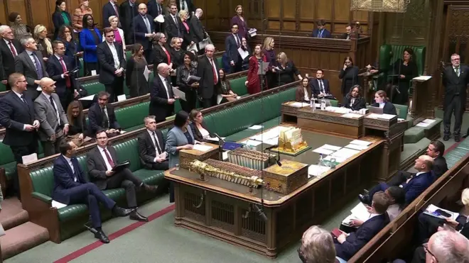 イギリス下院の本会議室。与党閣僚と議員たちが議長に向かって左側のベンチ席に座ったり、立ったりしている。中央の大きい演説用テーブルに置かれた「ディスパッチ・ボックス（公文書箱）」の前では、ブルーグレイの上着を着たマムード内相が立ち、箱に書類を置いて話している。立っている与党議員たちは、立席することで内相発言への指示を表している。手前には保守党など野党議員が、反対側の与党と同じ、緑の革張りのベンチ席に座っている。