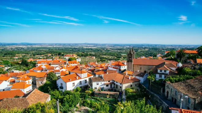 Vista do município de Fundão, em Portugal, com casas brancas de telhados de telha laranja e uma igreja, num vale cercado de árvores, e num dia de céu azul