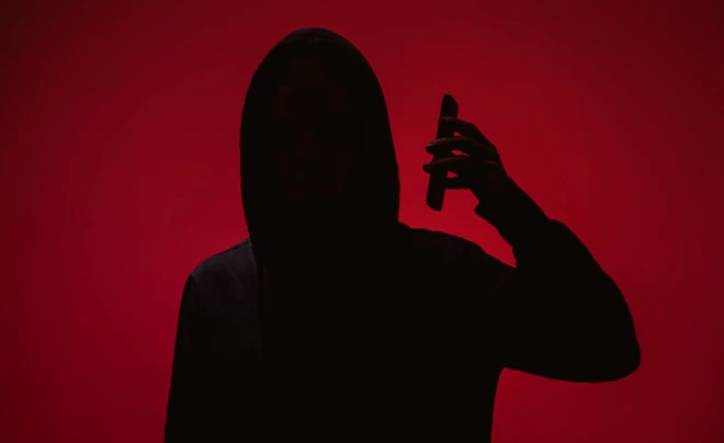 ظل شخص يرتدي قبعة رأس ويمسك هاتفاً محمولاً قرب أذنه، بخلفية حمراء موحّدة، مع إخفاء كامل لملامح الوجه 