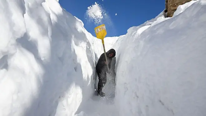 Bir kişi montlu kapşonu kapalı elinde kürekle boyundan yüksek karların arasından açtığı yoldan kazarak ilerliyor.