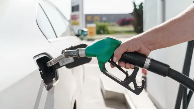 Рука людини тримає паливний пістолет із зеленою ручкою та заправляє білий автомобіль.