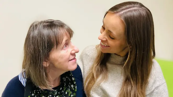 Patient Samantha Denison is sitting on a hospital bed and looking at her daughter Steph. Samantha has shoulder length brown hair and wears a dark blue cardigan over a floral top. Steph has long brown hair and wears a light grey, high-neck jumper.