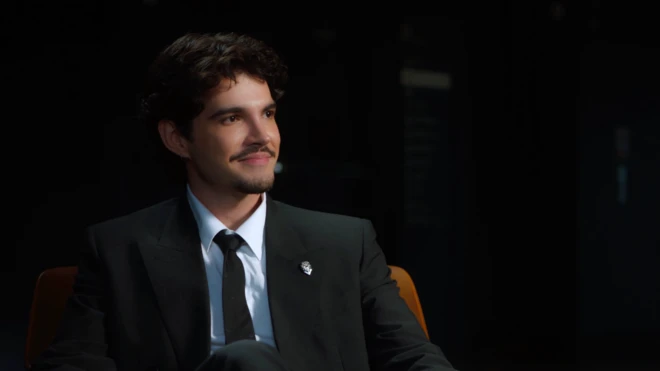 Jão, de cabelos escuros e bigode, veste terno preto, camisa branca e gravata preta, enquanto está sentado em uma poltrona. Ele sorri discretamente e olha para fora do enquadramento, em um ambiente escuro iluminado por luz direcionada.