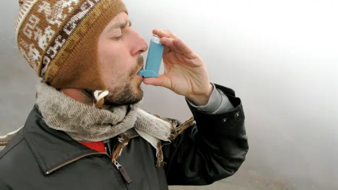 Un homme utilise un inhalateur bleu lors d'une promenade brumeuse. Il est chaudement emmitouflé dans un chapeau marron, une écharpe grise et un manteau noir.