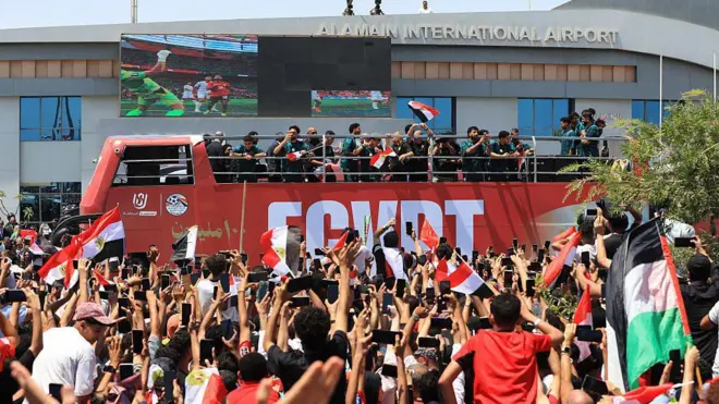 لاعبو المنتخب المصري يلوحون بأيديهم من على متن حافلة مكشوفة، وسط حشود من المشجعين الذين تجمعوا لاستقبالهم في مطار العلمين الدولي بمدينة العلمين الساحلية الشمالية في 10 يوليو/تموظ عام 2026. ويظهر مشجعون يلوحون للاعبين ويحملون أعلام مصرية، ويحاول بعضهم التقاط صور بهواتفهم المحمولة.