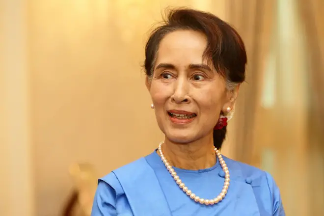 အပြာရောင် ချည်ရင်ဖုံးအင်္ကျီ၊ ပုလဲဆွဲကြိုးနဲ့ ပန်းအနီရောင်ပန်ပြီး ပြုံးနေတဲ့ ဒေါ်အောင်ဆန်းစုကြည်