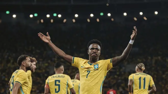 Vini Jr., com as mãos para o alto, acompanhado de outros jogadores da seleção brasileira de costas, todos uniformizados, em um estádio de futebol lotado