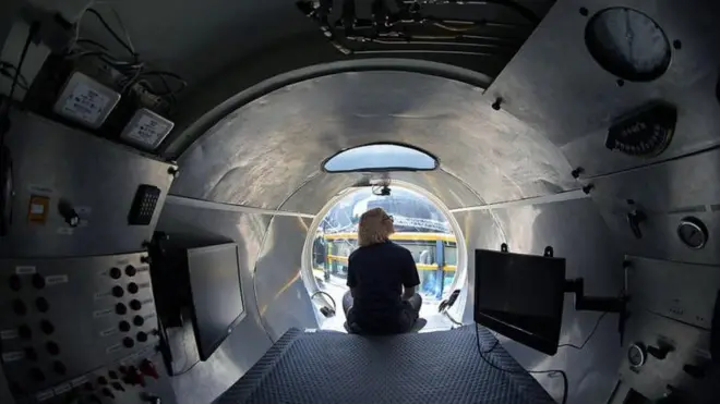 Vista interna do submarino, com mulher sentadasudeste esportecostas na ponta