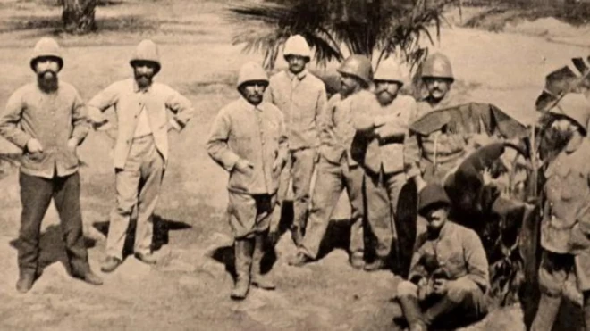 Ancient French colonial masters wear boots and oversize coats dey pose for a photo