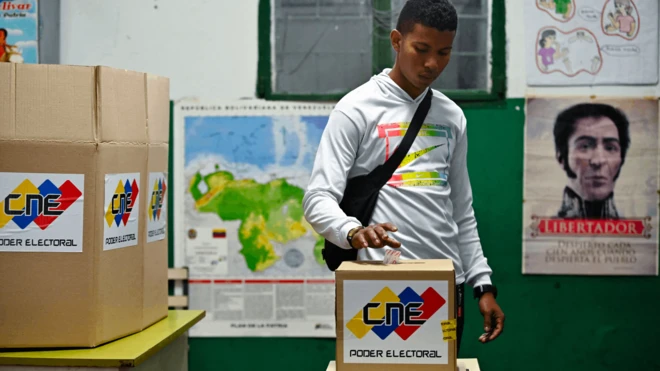 Un hombre con una camiseta blanca vota en una urna que dice "CNE Poder Electoral".