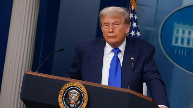 Donald Trump mu mwambaro wirabura n'ikaruvati y'ubururu ahagaze ahakirurutse muri White House mu cumba yakiriramwo abanyamakuru 