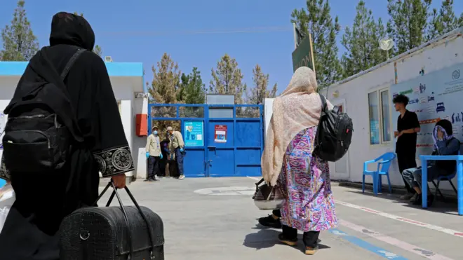 تصویر دو زن بازگشته در یکی از مراکز پذیرش مهاجران وابسته به ملل متحد. این زنان با اندک وسایل دست داشته شان که شامل یک بیک میشود راهی افغانستان شدهاند. یکی از این زنان یک دیک بخار نیز به دستش است. در تصویر چند مأمور کارمند در این مرکز نیز به چشم میخورند.