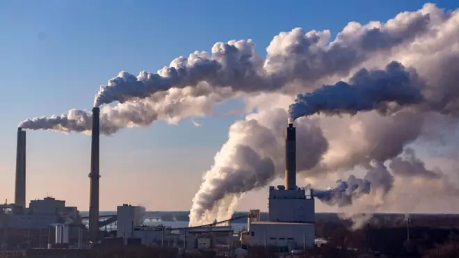 Wind is blowing clouds of pollution from the large smoke stacks of coal burning power plant. It is daytime and the sky is otherwise clear and bright blue.