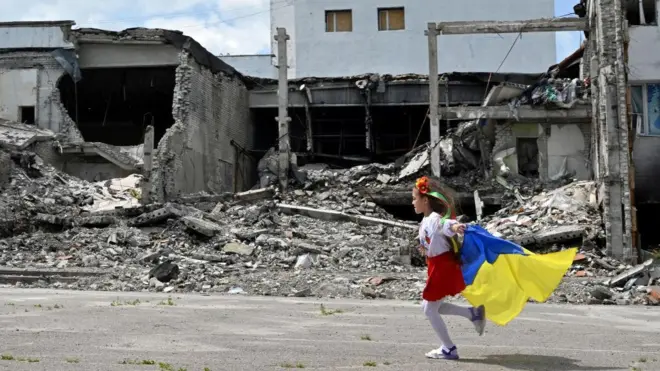 A child runs as she holds a Ukranian flag in front of the destroyed building of a cultural centre, hit in a missile strike, during a graduation ceremony of art students, in the town of Derhachi, Kharkiv region, on June 17, 2023.