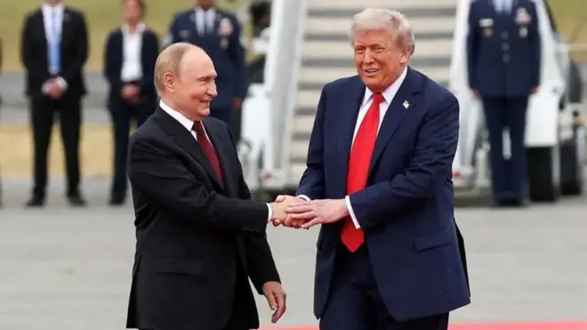 Russian President, [left] Vladimir Putin wear black coat, white shirt and maroon red tie. US President, [right] Donald Trump wear navy blue suit, white shirt and pepper red tie. E dey shake hands wit Vladimir Putin and pipo wey dia face no show dey for di background.