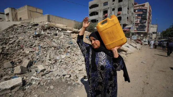 Palestinian woman carries a container of water as she passes by rubble in Gaza City (03/07/25)