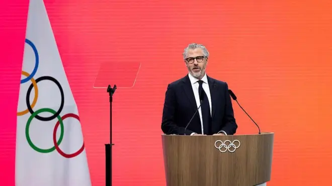 ダークスーツ姿のワッサーマン氏が、五輪マークがついた演台に立ち口を開いている。近くには五輪旗が立てられている