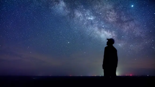 Gece yıldızlı gökyüzüne bakan şapkalı bir adam. 