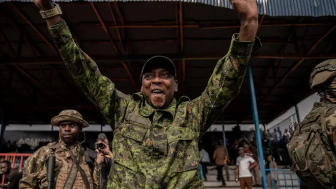 Lt Col Willy Ngoma yambaye imyenda ya gisirikare azamuye amaboko arimo guseka asuhuza abantu. Iruhande rwe hari abasirikare babiri bamurinze. Inyuma hari abandi bantu benshi. Ifoto yo ku itariki ya 6 Gashyantare (2) mu 2025, i Goma, ku kibuga cya Stade de l'Unité.