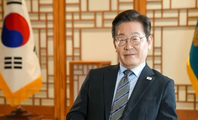 검은색 정장을 입은 이재명 대통령이 서울 집무실의 의자에 앉아 있다. 뒤로는 태극기와 대통령 표장기가 있다.