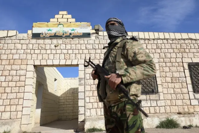 A Hayat Tahrir al-Sham fighter in military gear stands outside a building in Hama, Syria, bearing the Syrian flag and President Bashar al-Assad's face over the door.