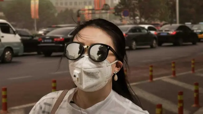 Seorang perempuan berjalan di trotoar kota sambil mengenakan kacamata dan masker.