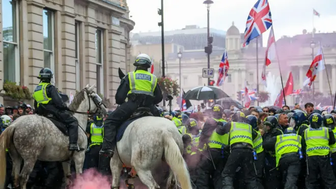 Конные и пешие полицейские в светоотражающих жилетах пытаются удержать толпу с флагами Англии и Соединенного Королевства на стыке Уайтхолла и Трафальгарской площади в Лондоне под дождем