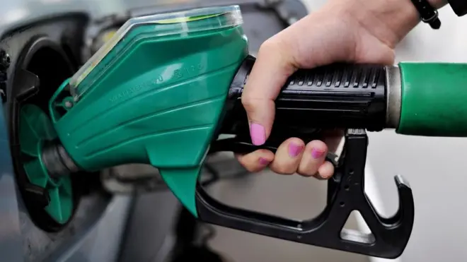 A close-up of a woman's hand, with pink nail varnish, holding a green petrol pump.