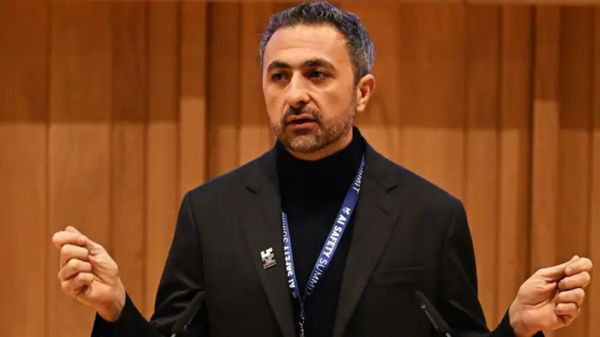 Mustafa Suleyman, vestido de negro y con un cordón de la cumbre de seguridad de la IA alrededor, se dirige al público.