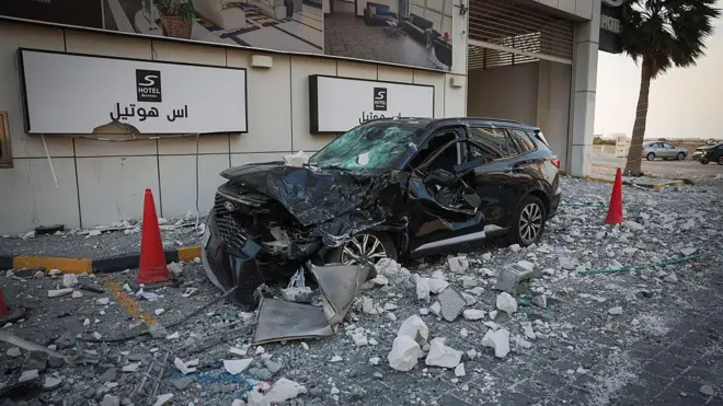 سيارة سوداء محطمة تقف بين مخروطين مروريين أحمرين وسط الأنقاض خارج فندق في المنامة، البحرين، يوم الأحد 1 مارس/آذار 2026، مع وجود سيارتين أخريين خلف شجرة نخيل في الخلفية.
