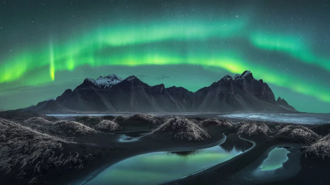 Here's a mountain on a volcanic black sand beach in Iceland. It's surrounded by large dunes - which are rounded piles of sand or small bumps created by the wind. The sky is completely covered by the Northern Lights. Beautiful!