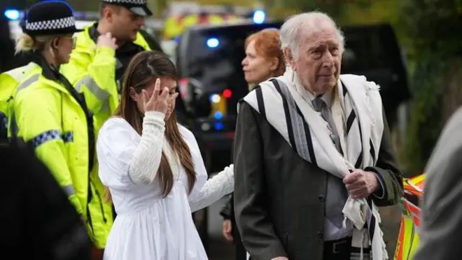 رجل يرتدي زي يهودي وخلفه سيدة ورجال شرطة يقفون خلفهما امام كنيس هيتون بارك في مانشستر