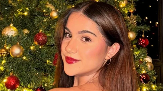 A woman looks back over her shoulder, wearing red lipstick and gold hoops, in front of a Christmas tree