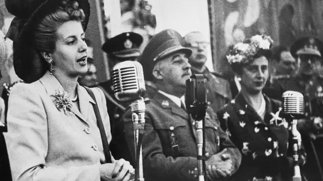 Eva Perón habla junto a un micrófono durante su visita a España en 1947. A su izquierda, de pie como ella, el general Franco y su mujer, Carmen Polo.