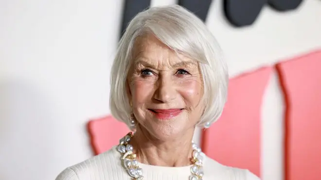 Helen Mirren ma białe włosy przycięte na boba i patrzy w prawo od aparatu. Ma na sobie perłowe kolczyki i gruby naszyjnik ze złota i pereł, a na sobie biały top.