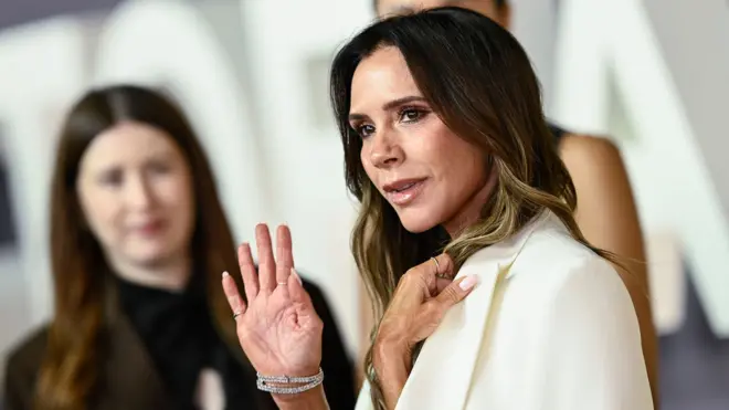 Victoria Beckham waves while wearing a white suit with other people in the background as she attends the Victoria Beckham premiere in London on Wednesday.