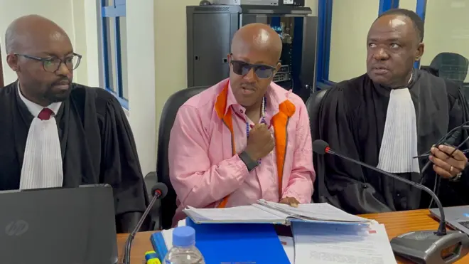 Aimable Karasira (yicaye hagati) akoze ikimenyetso cy'igipfunsi mu gatuza, arimo kuvuga. Yambaye amashapule abiri, isaha, indorerwamo z'umukara n'imyenda y'iroza iranga imfungwa. Mu kiganza kimwe afashe ikayi nini ifunguye. Akikijwe n'abanyamategeko babiri bamwunganira. Ifoto yo ku wa gatatu, ku itariki ya 28 Gicurasi (5) mu mwaka wa 2025. 