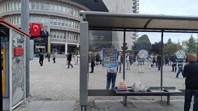 Otobüs durağı ve arkada belediye binası var. Çok sayıda kişi geniş bir alanda telefonla konuşuyor