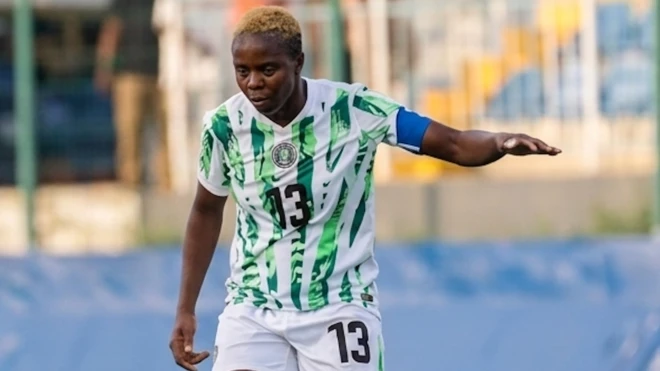 Ngozi Okobi-Okeoghene arbore des cheveux courts teints en blond et porte un maillot de football nigérian blanc et vert ainsi qu'un short blanc floqué du numéro 13. Son bras gauche est tendu sur le côté pour garder l'équilibre tandis qu'elle regarde attentivement le terrain pendant un match de football. À l'arrière-plan, on aperçoit, de façon floue, des barrières placées devant une tribune.